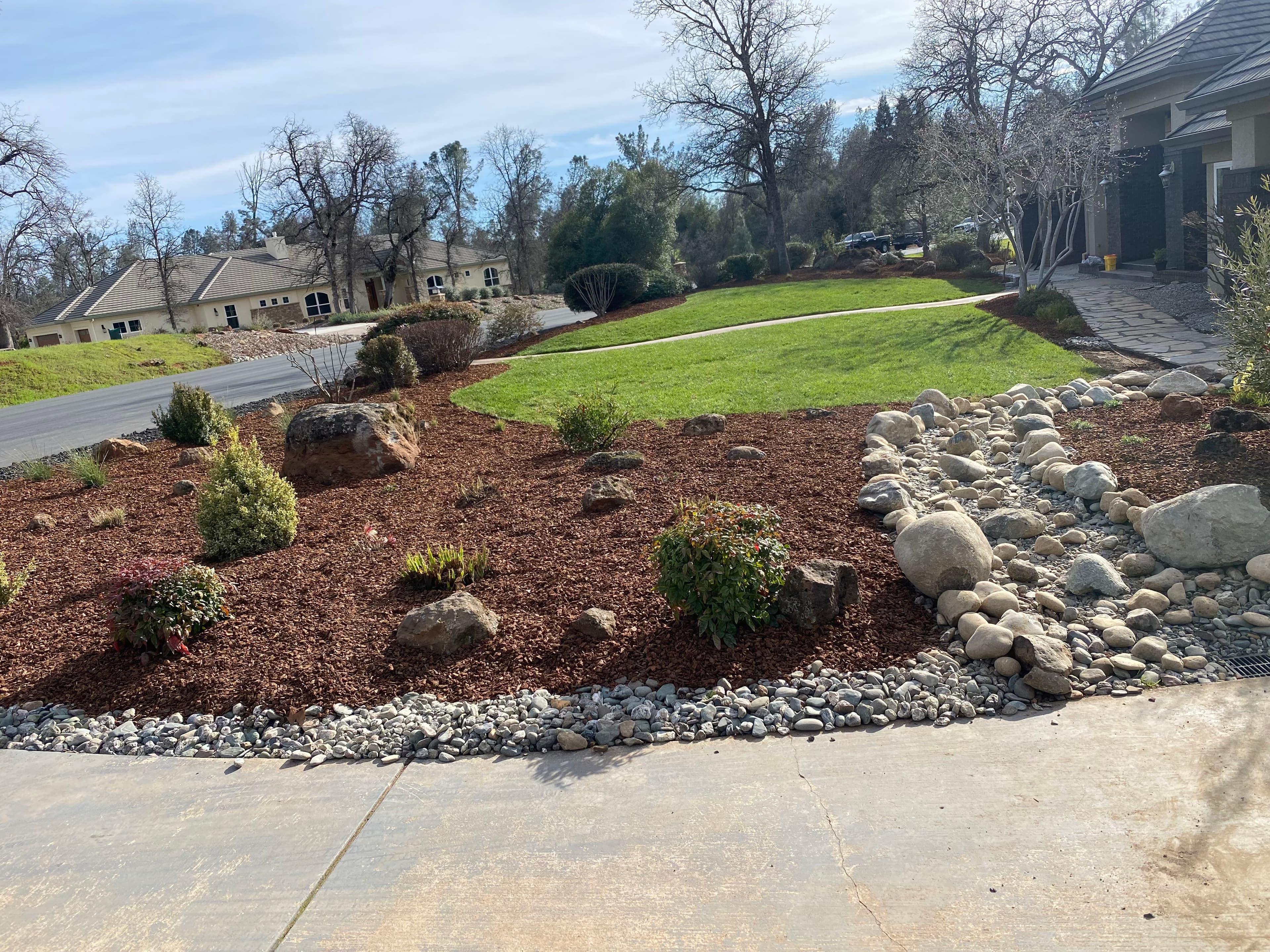 Landscaped yard with brown mulch, small shrubs, a dry rock creek bed, and green lawn.