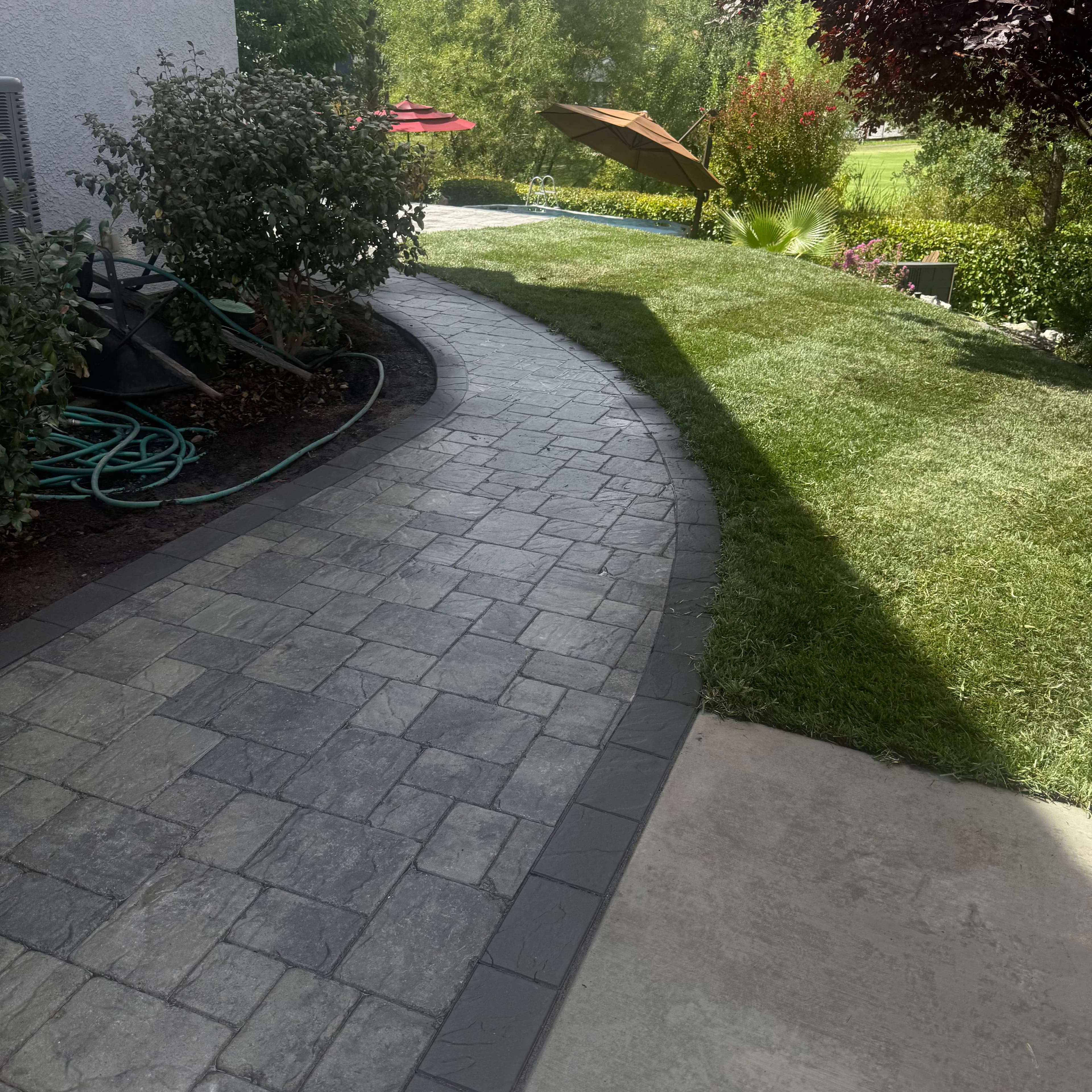 Curved grey stone paver walkway winding through a lush green lawn in a sunny backyard.