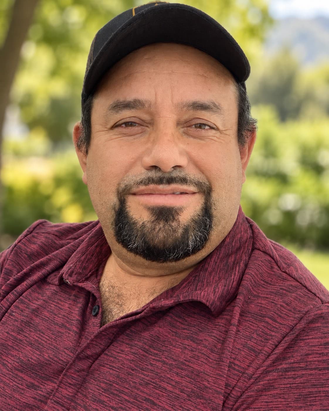 Smiling man with a goatee and black baseball cap wearing a maroon polo shirt outdoors.