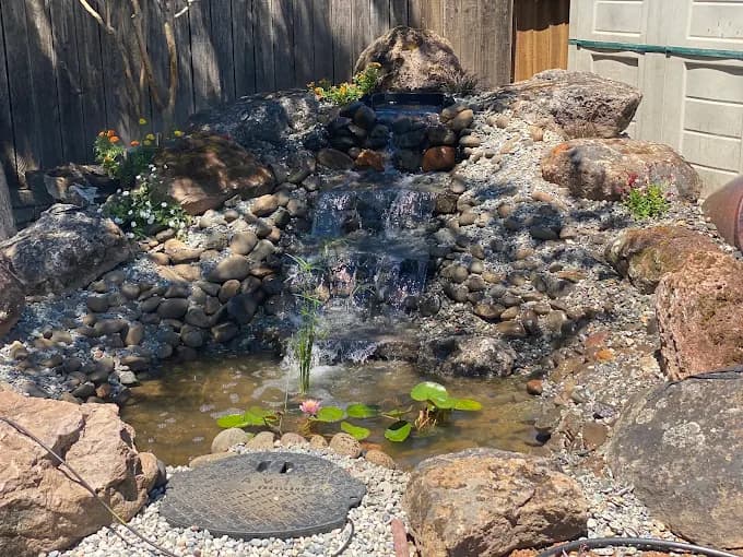 Tiered rock waterfall cascading into a garden pond with lily pads and surrounding decorative boulders.